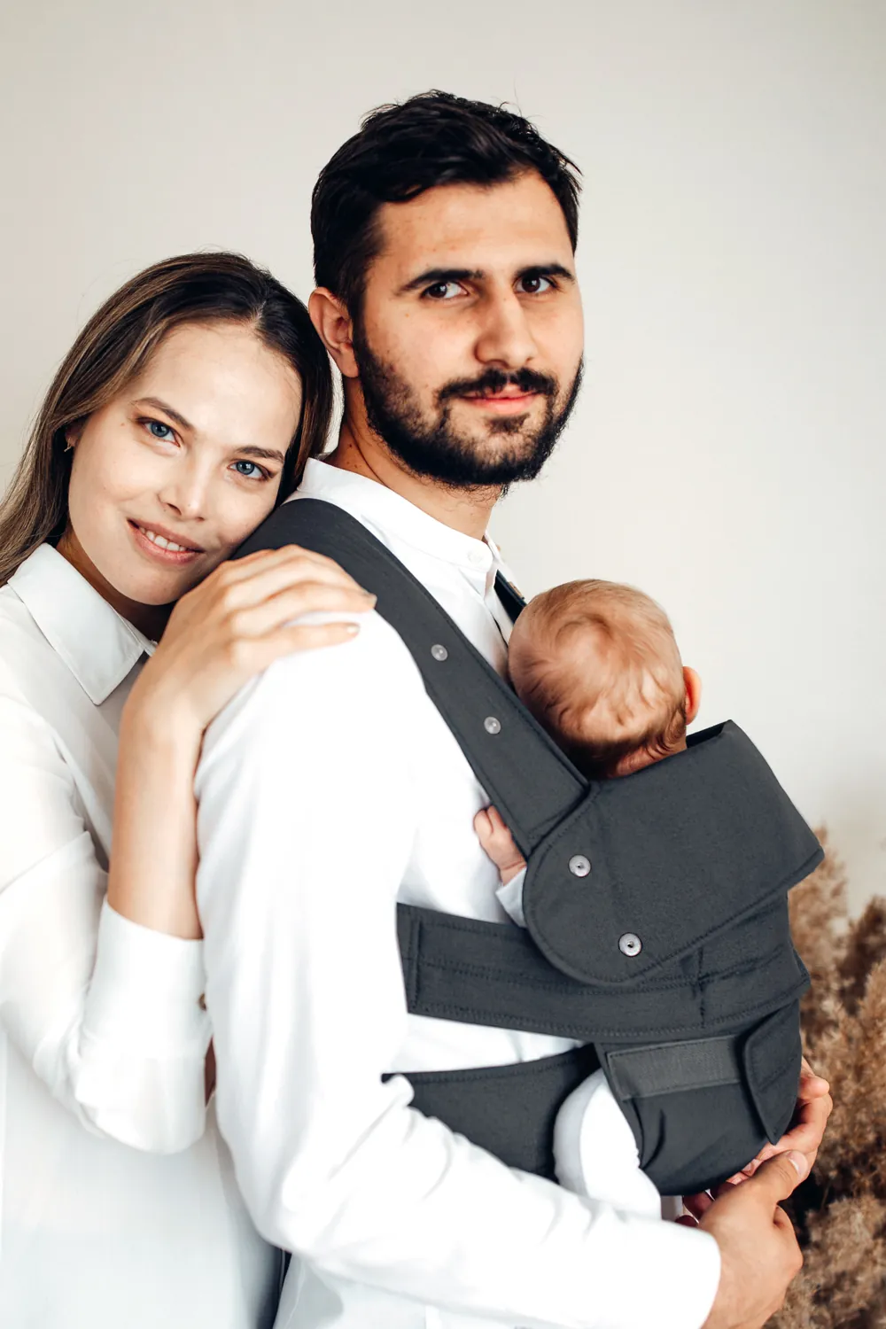 Paarfoto: Mutter umarmt den Vater, der ein Baby in der Marsupi Classic 2 Babytrage in Grau trägt – Portraitaufnahme.