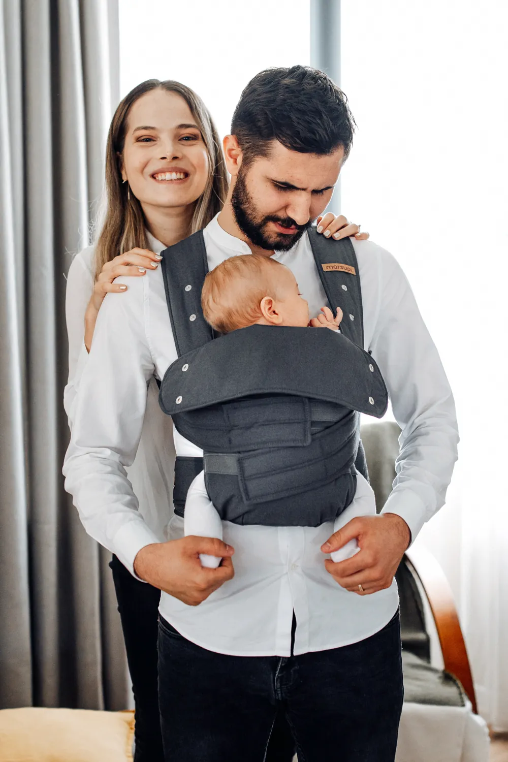 Familienfoto: Mutter steht hinter dem Vater, der ein Baby in der Marsupi Classic 2 Babytrage in Grau trägt – Indoor-Aufnahme.