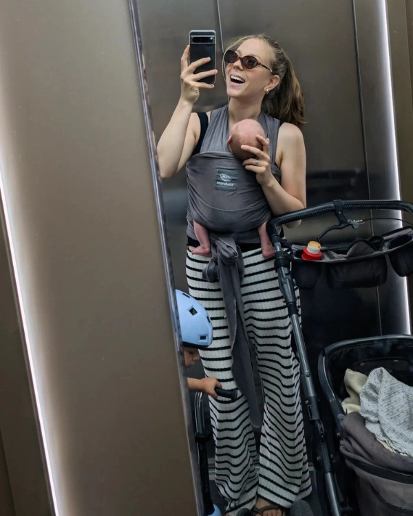Spiegel-Selfie einer lachenden Frau, die ihr Baby in einer dunklen manduca Babytrage trägt und lässig gestreifte Hosen anhat.