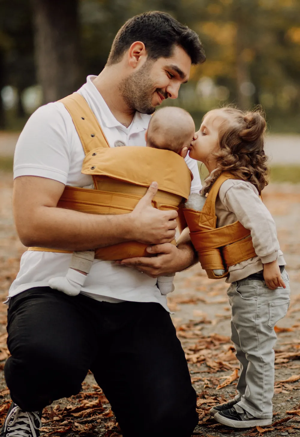 Kleinkind küsst Baby, Papa trägt Baby im Marsupi Classic 2 Butterscotch im Park