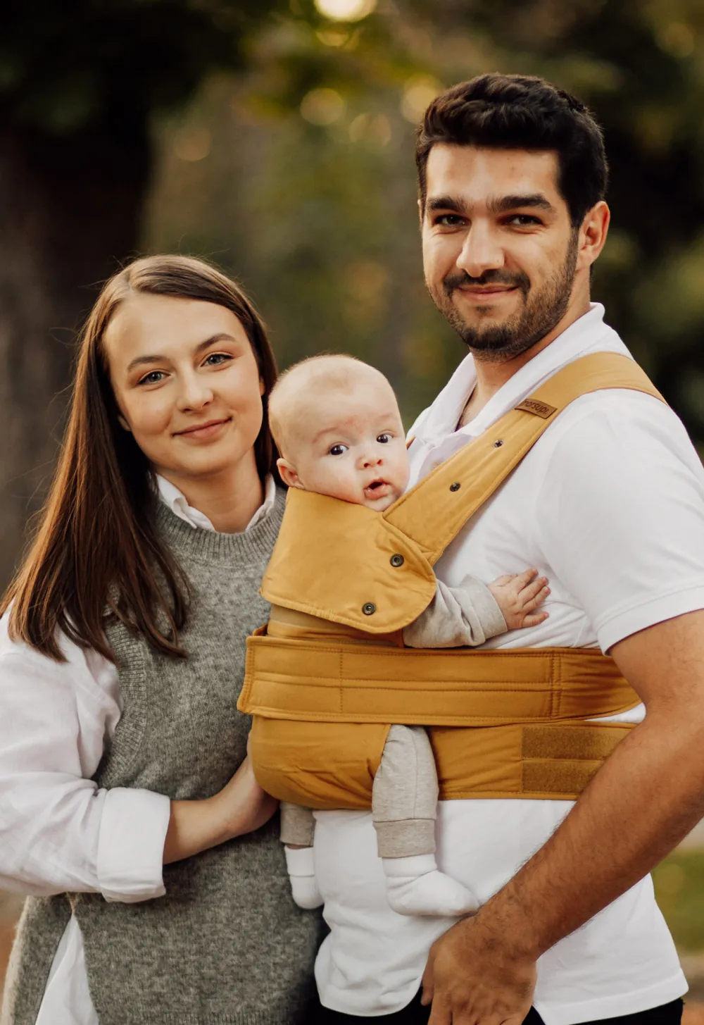 Eltern stehen im Park, Papa trägt Baby im Marsupi Classic 2 Butterscotch und beide schauen zur Kamera