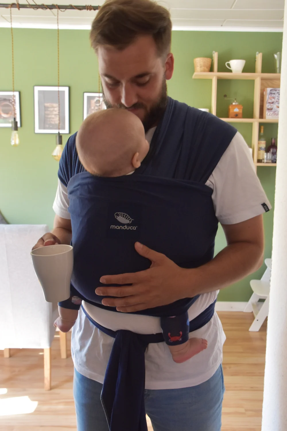 Mann trägt Baby vorne im manduca Sling Babytragetuch Navy in der Küche