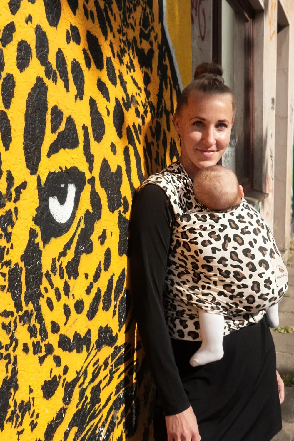 Frau mit Baby im manduca Sling baby-tragetuch im Leo-Design vor gelber Leopard-Wandmalerei