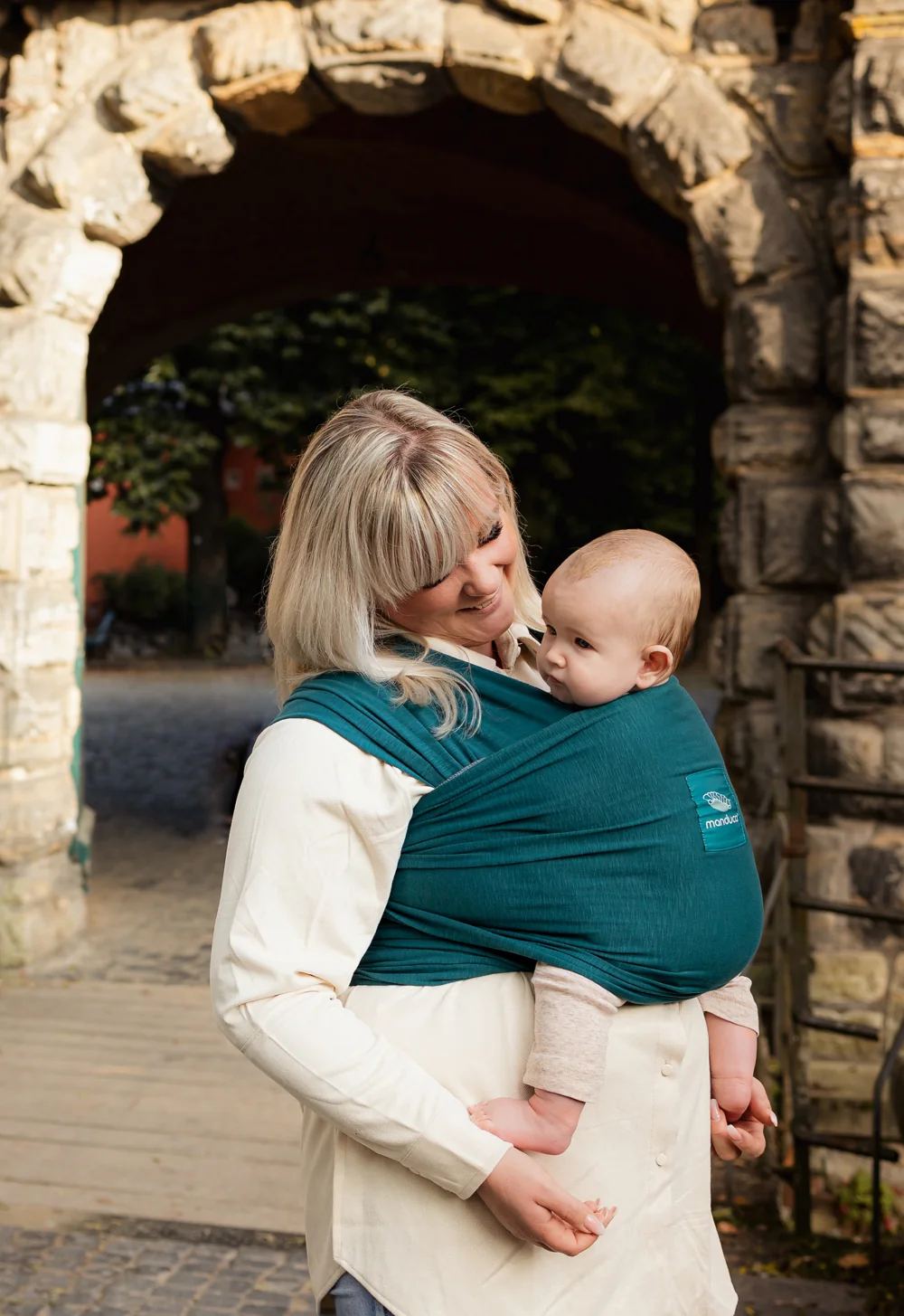 Frau trägt Baby im manduca Sling Babytragetuch Teal vor einem Steinbogen