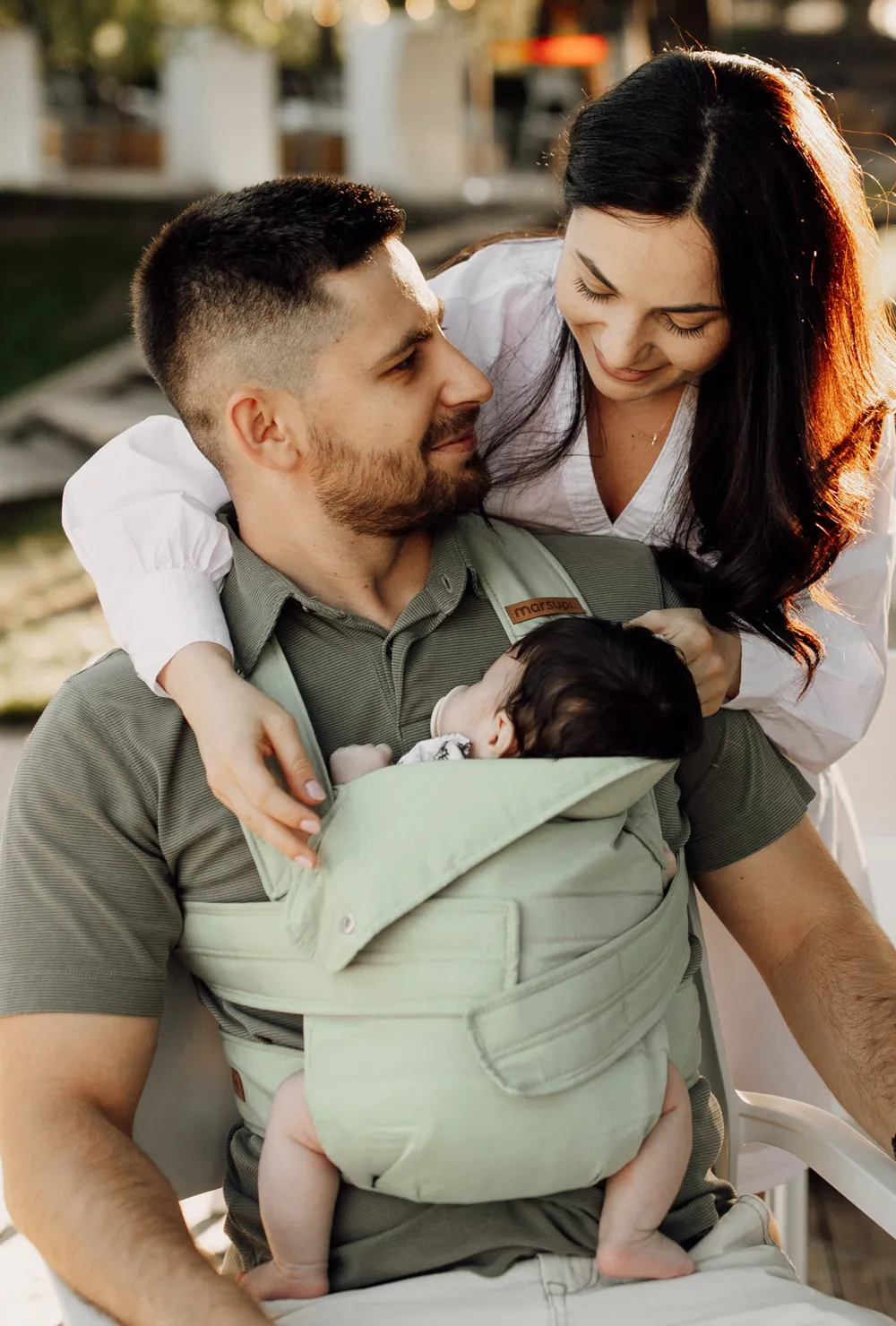 Paar: Vater trägt Baby in der Marsupi Breeze Babytrage Pistachio, Mutter hält das Baby – Lifestylefoto