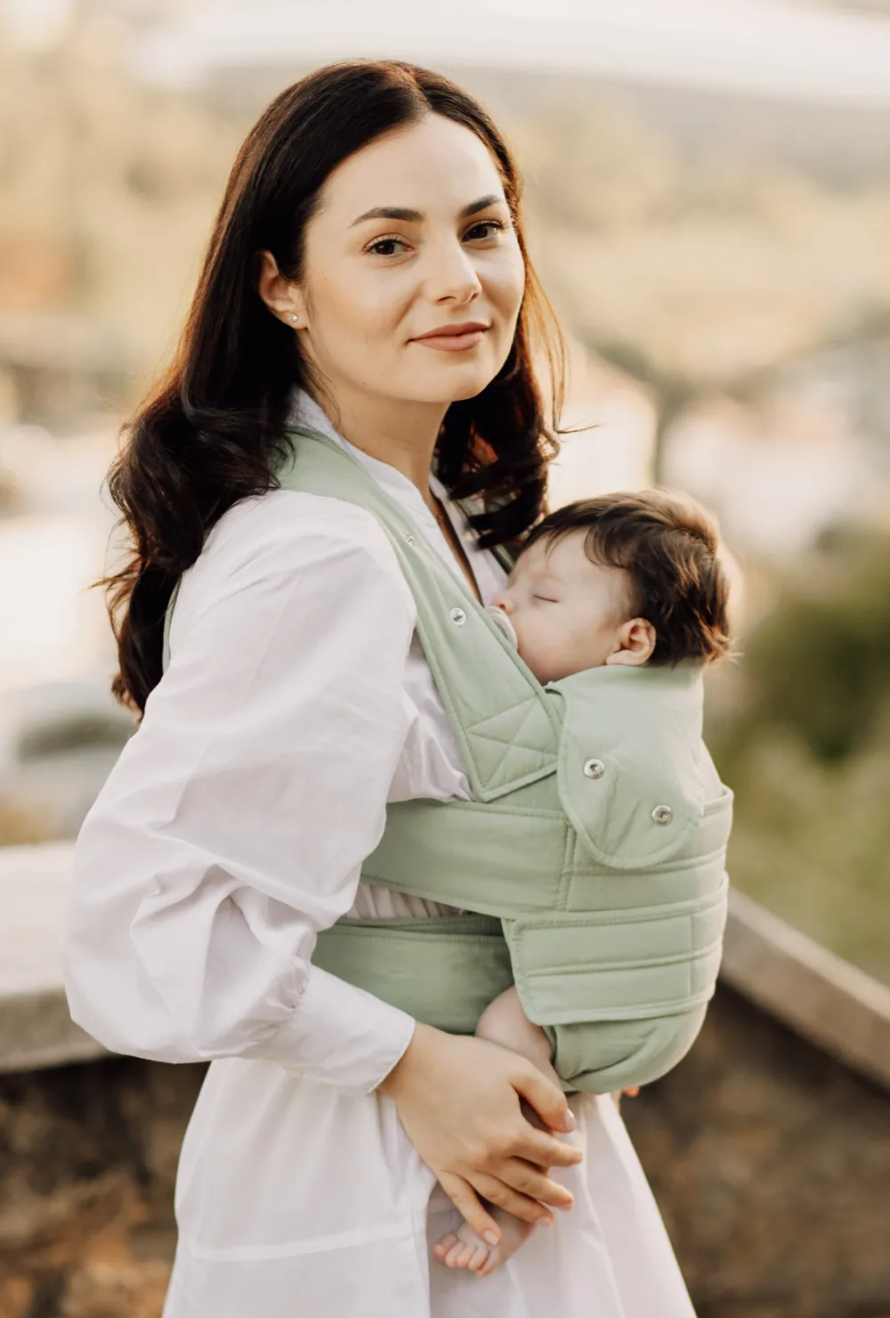 Frau blickt in die Kamera, Baby schläft in der Marsupi Breeze Babytrage Pistachio – Outdoor