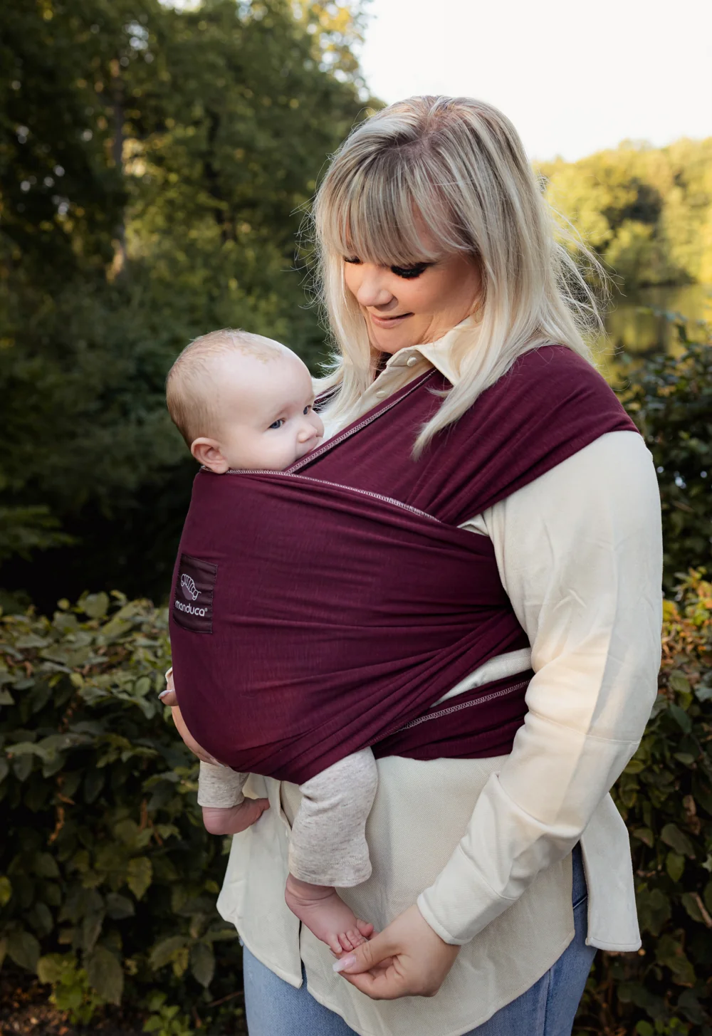 Frau trägt Baby im manduca Sling Babytragetuch Berry vor grüner Naturkulisse