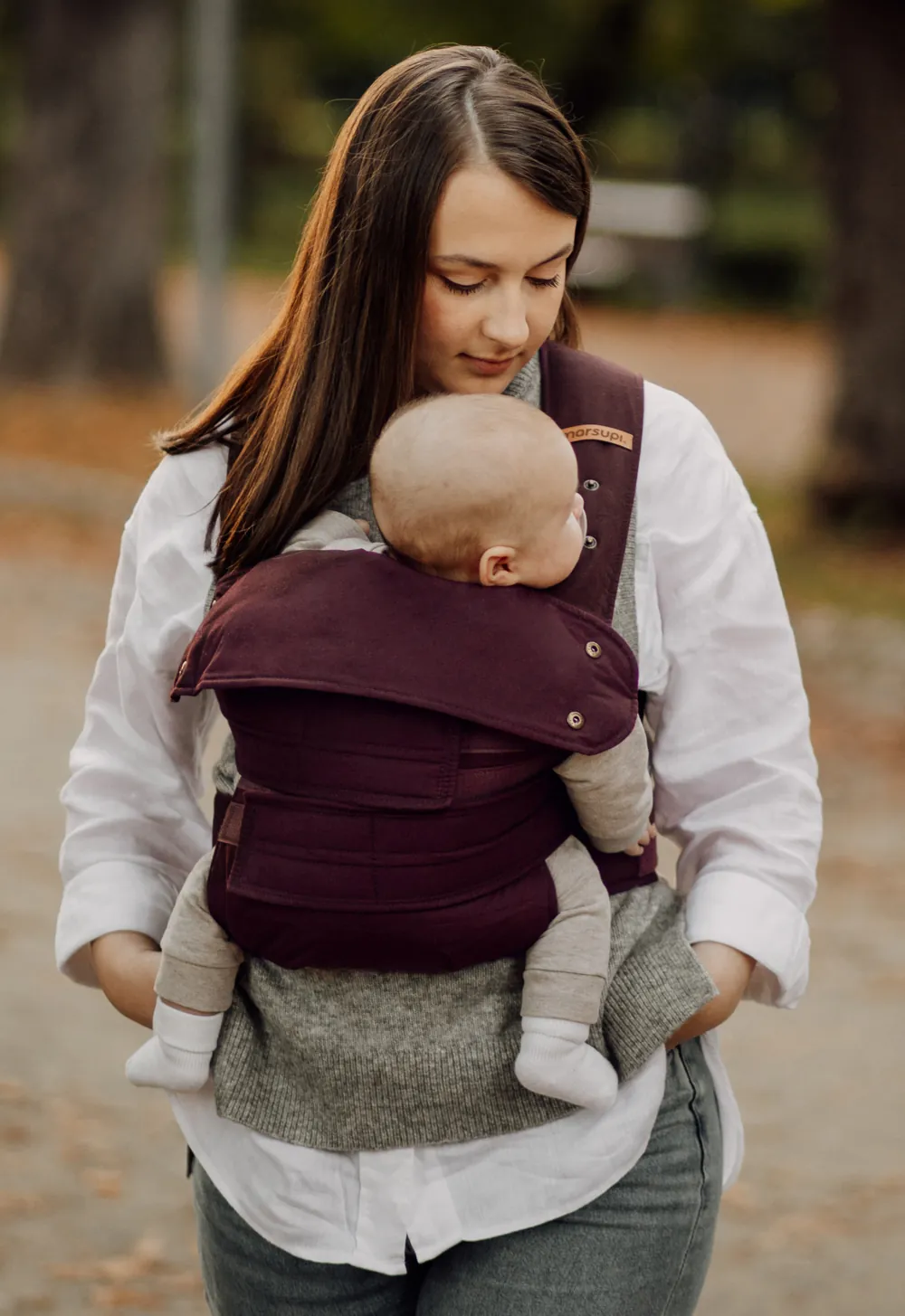 Mutter trägt Baby in Marsupi Classic 2 Babytrage Berry und hält ein Kleinkind an der Hand im Park