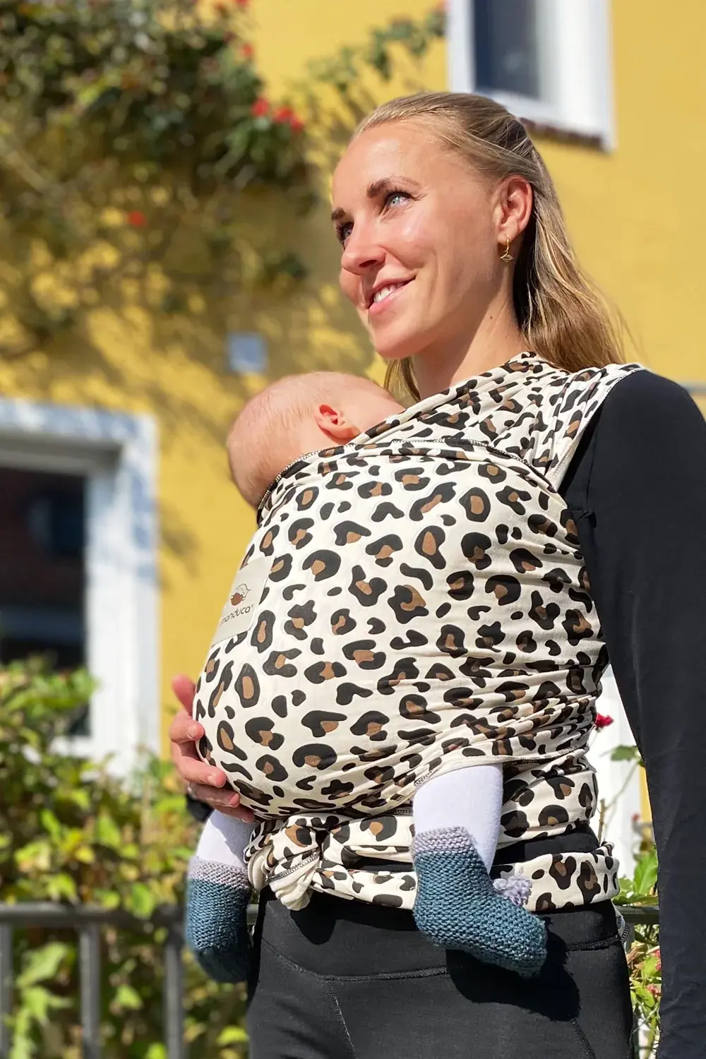 Seitliche Ansicht: Frau trägt Baby im manduca Sling baby-tragetuch im Leo-Design vor gelbem Haus